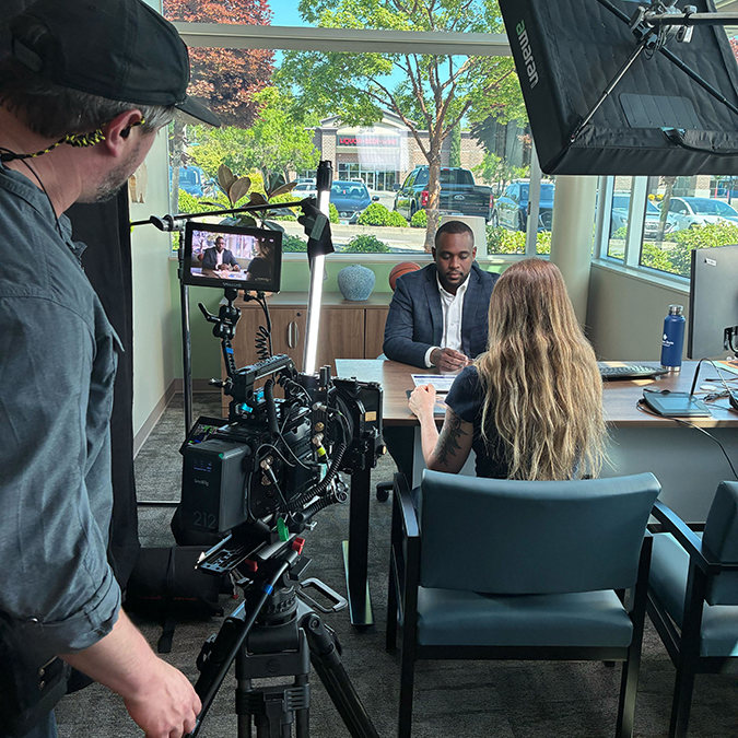 A film crew shooting a commercial inside a credit union branch, with a camera operator and lighting equipment in the foreground filming two people seated at a table.