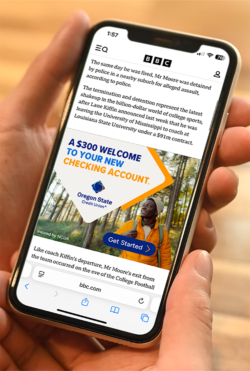 A close-up of a hand holding a smartphone displaying an Oregon State Credit Union mobile ad offering a $300 welcome bonus for a new checking account, overlaid on a news article in a mobile browser.